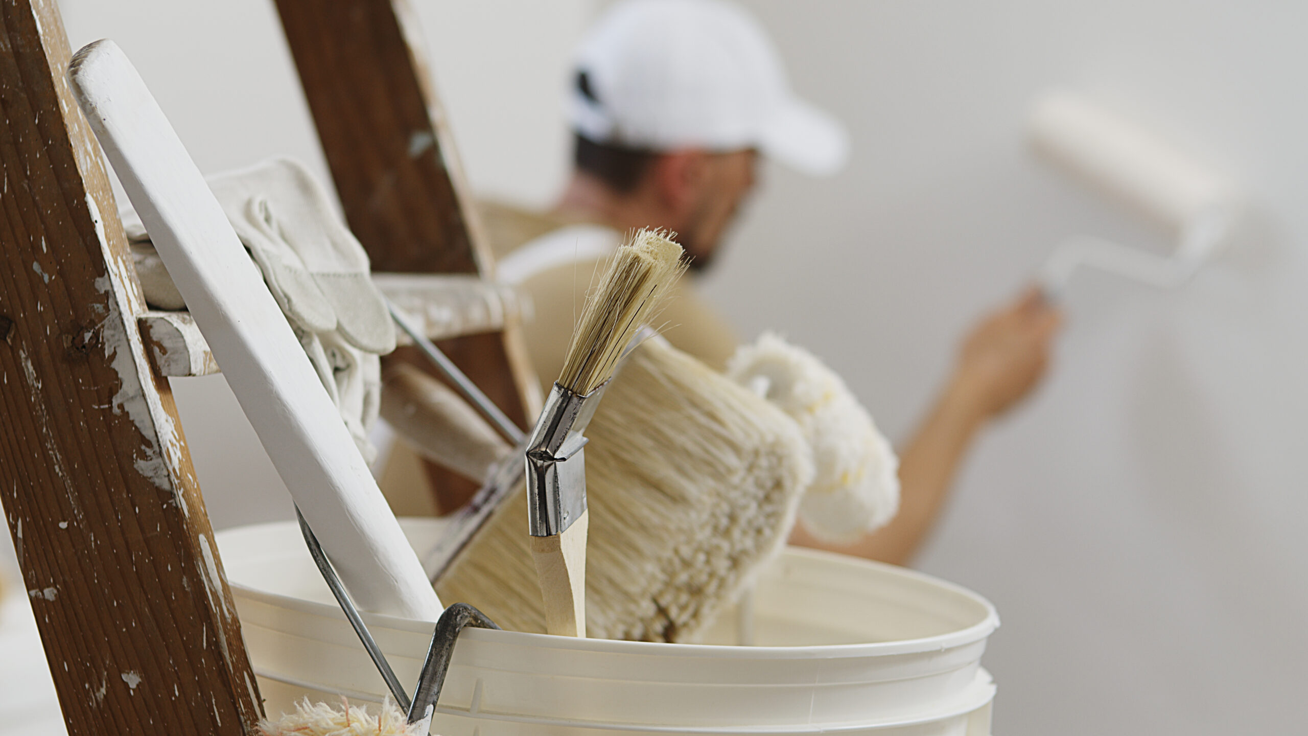 hands of house painter man decorator work of home to renovate, using roller paint, white bucket on wooden ladder with paint brushes as background, close-up